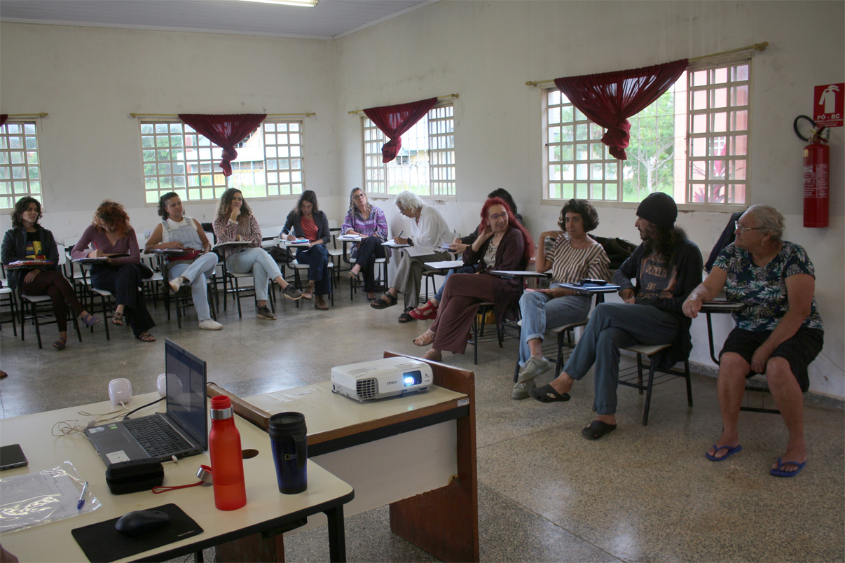 Turma de estudantes de Audiodescrição. Foto da frente da sala de aula onde se pode ver estudantes sentades em carteiras ao redor da sala, assistindo aula. À direita em primeiro lugar está D. Belinha, aluna com baixa visão e em formação para atuar como consultora.