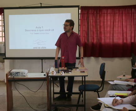 Primeiro dia de aula: A fotografia do fundo da sala de aula uma tela projeta uma frase: “Descreva o que você vê”.
                  Ao lado da tela uma cortina vermelha cobre a janela.
                  À frente o professor Tabuh um homem branco com cabelos pretos e curtos, usando óculos, veste camisa vermelha e está em pé atrás de uma mesa onde está o projetor.
                  À esquerda dele estão carteiras com pessoas sentadas.