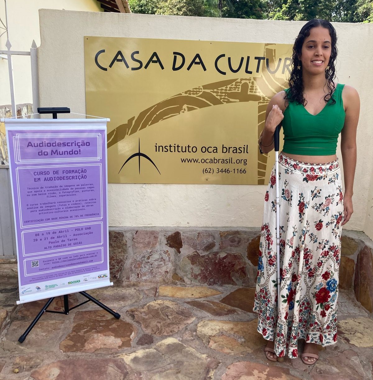 Viviane Santos Queiroz, professora do Curso Audiodescrição do Mundo!, em frente à Casa de Cultura Oca Brasil onde  estava a Exposição 'Corpo Segredo, adentrando Corpos e histórias'.  Alunes do curso interagiram fazendo roteiros de descrição das obras e lendo-os em voz alta enquanto ela tocava as peças de arte textil e bordados, desdobramentos das fotografias.