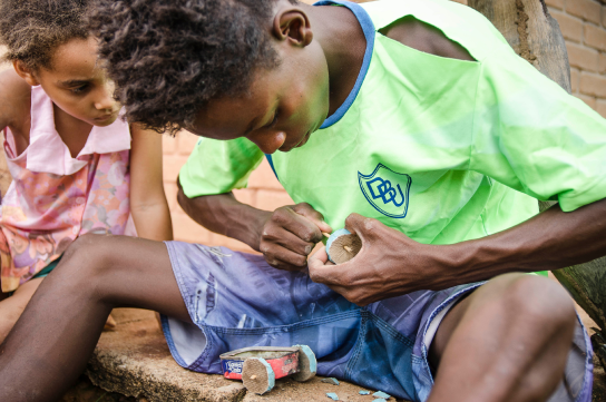 Um adolescente de pele negra veste bermuda azul e camiseta verde e monta um carrinho com lata de sardinha e rodas de borracha de sandálias havaianas. A seu lado uma menina acompanha atenta.