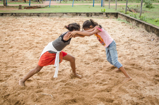 Duas meninas brincam de luta em um campo de areia. Se seguram pelos ombros para desequilibrar a outra