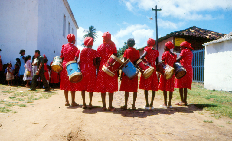 Sete Caixeiras, vestindo vermelho, estão com suas caixas penduradas no ombro e esperam, do lado externo da Igreja do Carmo, em Alcântara, pelo Império que assiste à missa no domingo do Festejo. 
                  Em Alcântara, não é permitida a presença delas com seus instrumentos na igreja. 
                  Foto de Juvenal Pereira. Década de 1980.
                  